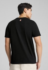 Sorte bomulds t-shirt med korte ærmer, der har et lille farverigt logo ved nakken på bagsiden. Glat tekstur, almindelig pasform.