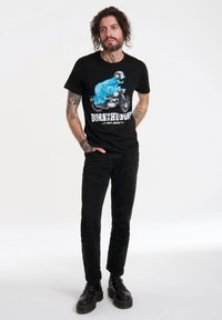 Camiseta negra con un gráfico azul de un personaje en una motocicleta y el texto "NACIDO PARA TENER HAMBRE" debajo, combinada con pantalones negros y botas.