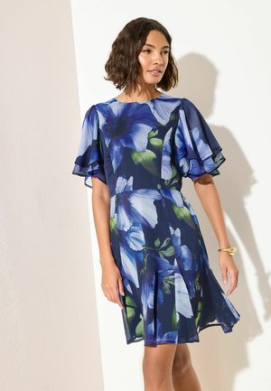 Femme aux cheveux bouclés mi-longs portant une robe bleu marine à fleurs avec des manches courtes superposées, debout contre un mur blanc.