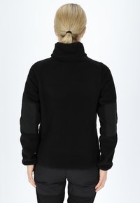 Giacca in pile nera con colletto alto, tessuto con texture e dettagli neri su spalle e gomiti. Vestibilità aderente con maniche lunghe.