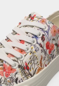 Κοντινό πλάνο σε άσπρο αθλητικό παπούτσι με άσπρα κορδόνια και λουλουδάτο μοτίβο σε κόκκινο, ροζ, κίτρινο και μοβ σε καμβαδένιο ύφασμα.