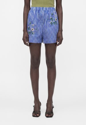 Frau trägt blau-weiß vertikal gestreifte Shorts mit rosa Blumenstickerei, kombiniert mit schwarzen Riemchenabsätzen und einem olivgrünen Top.