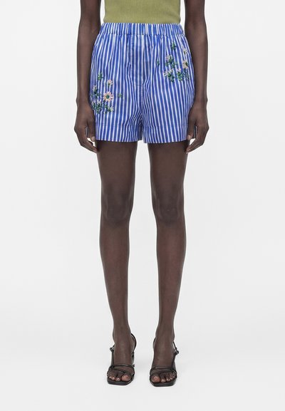 Femme portant un short rayé bleu et blanc vertical avec une broderie florale rose, associé à des talons noirs à lanières et un haut vert olive.