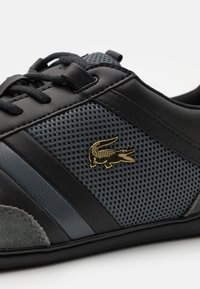 Černé a šedé tenisky s síťovými panely, černými šňůrkami a malým zlatým logem krokodýla na boku.