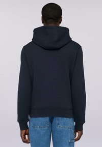 Námořnická mikina s velkou kapucí na šňůrku a vroubkovanými manžetami, v kombinaci se světle modrými džíny, pohled zezadu.