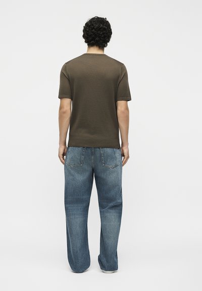 Άνδρας με σγουρά μαλλιά, που φοράει απλό καφέ κοντομάνικο πουκάμισο και φαρδιά μπλε τζιν, βλέποντας από πίσω σε λευκό φόντο.