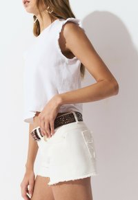 Top court blanc avec des manches froncées associé à un short en denim blanc. Ceinture marron avec des clous métalliques et un design texturé.