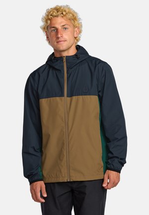 Chaqueta tipo rompevientos con capucha y mangas en azul marino, parte delantera en color tostado y detalles en teal a los lados. Ligera, con una textura suave.