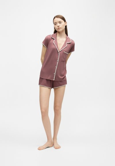 Jeune femme debout pieds nus portant un haut de pyjama mauve à manches courtes boutonné et un short assorti bordé de blanc sur un fond uni.