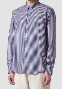 Hombre que lleva una camisa de botones azul y blanca a rayas verticales con un pequeño emblema de abeja dorada en el pecho y pantalones color crema.