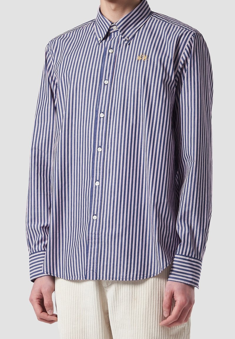 Mężczyzna w niebiesko-białą pionowo paskowaną koszulę z guzikami, z małym złotym emblematem pszczoły na piersi, oraz w kremowych spodniach.