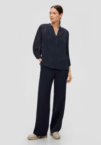 Marineblauwe geplooide blouse met een V-hals en lange mouwen, gecombineerd met bijpassende wijde broek en beige chunky schoenen. Glad, lichtgewicht stof.
