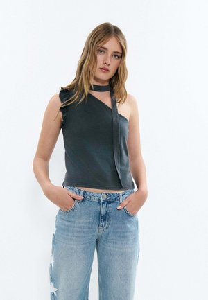 Jeune femme aux longs cheveux portant un haut sombre à une épaule avec détail choker et un jean bleu clair avec des étoiles sur les poches latérales.