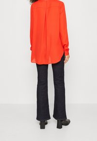 Blouse rouge à manches longues avec un pli dans le dos, associée à un pantalon évasé en denim foncé et des chaussures noires à talons blocs. Texture de tissu lisse.