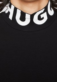 Černé tričko s vysokým límcem a bílým logem "HUGO". Hladká textura s přiléhavým střihem. Jednoduchá tmavá látka po celém povrchu.