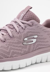 Lichtpaarse sportieve schoen met een gestructureerd mesh bovendeel, voorzien van een logo-accent en een witte tussenzool met een gekarteld zoolontwerp.
