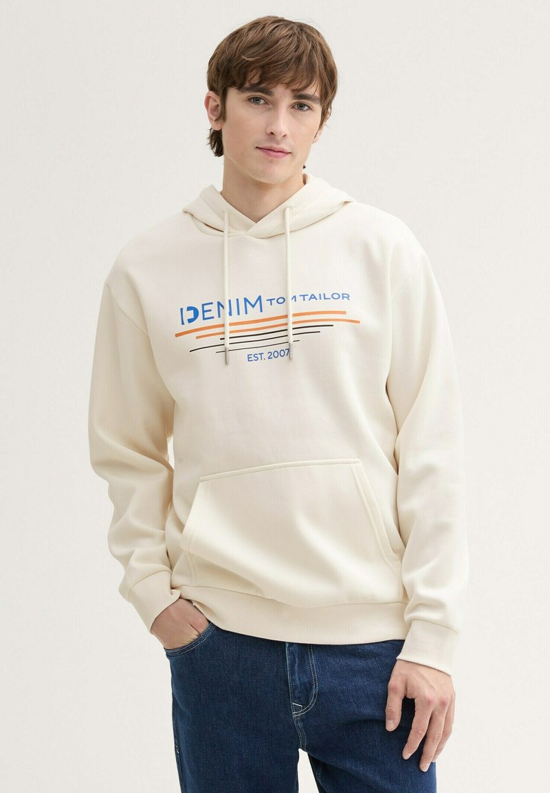 Crèmekleurige hoodie met een voorvak, voorzien van een blauw en oranje grafisch ontwerp, en een capuchon met trekkoord. Gekocht met een donkere spijkerbroek.