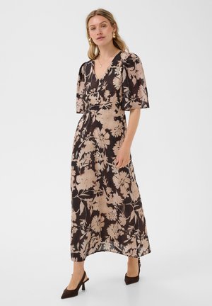 Femme portant une robe noire et beige à fleurs avec un décolleté en V, des manches jusqu'aux coudes et des chaussures noires à talons bas, debout devant un fond uni.