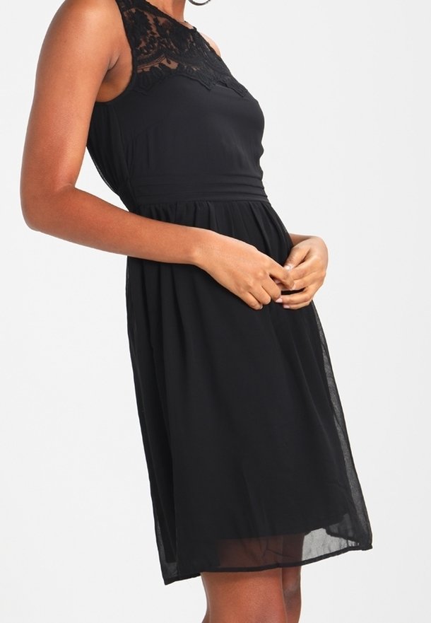 Femme portant une robe noire sans manches avec un détail en dentelle sur le haut de la poitrine et un tissu transparent superposé sur la jupe, debout les mains jointes.