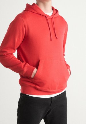 Person trägt einen roten Kapuzenpullover mit Fronttasche und schwarze Hose, Hände ruhen in der Tasche, auf einem einfarbigen hellen Hintergrund.