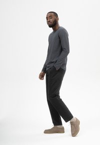 Langärmliges, dunkelgraues Hemd, schwarze Hose und hellbraune Wildlederschuhe mit Schnürung. Gerade Passform, glatte Textur und minimalistische Details.