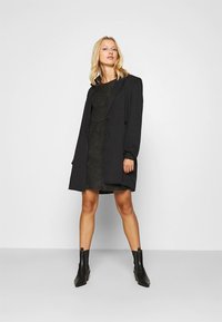 Crni kaput s dvostrukim kopčanjem, prevelikog kroja, glatke teksture i detaljima s gumbima, nošen preko tamne haljine teksture i gležnjača.