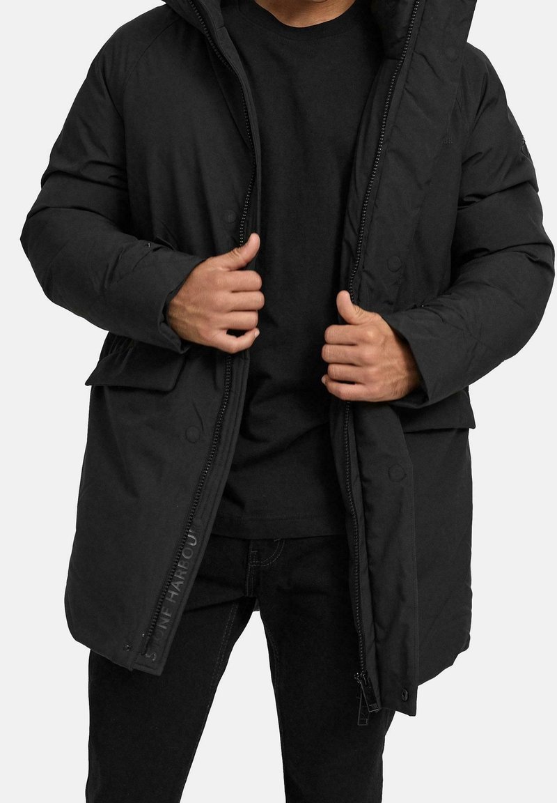Homme portant un manteau d'hiver matelassé noir par-dessus une chemise et un pantalon noirs, tenant le manteau ouvert au niveau de la poitrine, sur fond blanc.