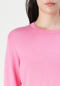 Roza pletena jopica s okroglim izrezom in dolgimi rokavi, ki se ponaša z gladko teksturo in diskretnim rebrastim robom na ovratniku in robovih.