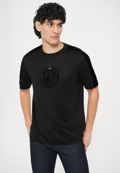 Fekete póló, középen egy texturált kerek logóval. A vállakon sima bársonyos díszítés található, amely fokozza az összhatást.