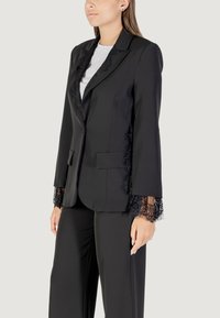 Blazer nero su misura con dettagli in pizzo lungo il colletto e i polsini, dal design strutturato e con tasche frontali. Abbinato a pantaloni coordinati.