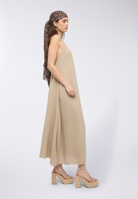 Abito maxi beige, fluido, con una texture increspata, abbinato a sandali con tacco alto e una sciarpa a stampa leopardata legata intorno alla testa.