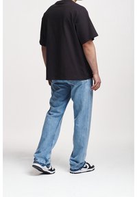 Camiseta negra de algodón con mangas cortas, combinada con jeans de mezclilla claros y zapatillas blanco y negro, vista desde atrás.