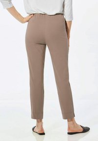 Pantalons marron à taille élastique, avec une coupe slim, jambes effilées et fentes aux chevilles. Le tissu a une texture lisse. Portés avec des chaussures noires à enfiler.