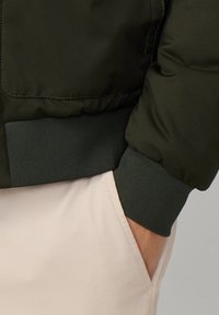 Donkergroene bomberjack met ribgebreide manchetten en tailleband. Gladde matte afwerking en subtiele stikseldetails. Roomkleurige broek met zichtbare zak.