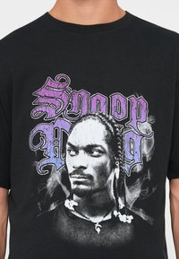 Sort bomulds t-shirt med en grafik af et ansigt med fletninger, omgivet af lilla og grå stiliseret tekst "Snoop Dogg."
