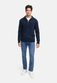 Mørkeblå strikket pullover med halvlukket krave, tekstureret stof og ribbede manchetter, parret med lyseblå jeans og hvide sneakers.