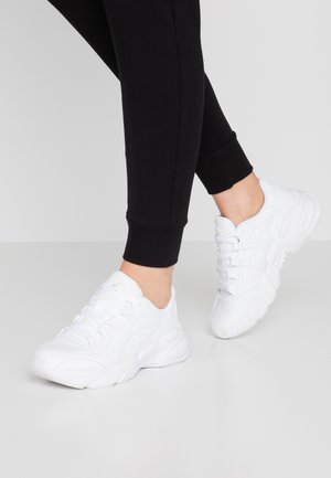 Personne portant des baskets blanches de sport et un pantalon noir ajusté, debout devant un fond gris clair uni.