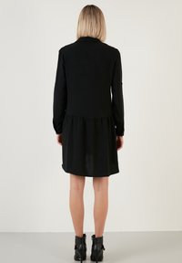 Vestito lungo nero a maniche lunghe con busto aderente e gonna a campana. Realizzato in tessuto testurizzato, con polsini con bottoni e scollatura alta.