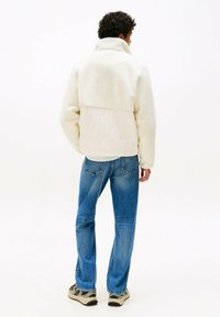 Fleece-Jacke in Creme mit einem gesteppte Unterteil, hohem Kragen und gerippten Bündchen, kombiniert mit blauen Jeans und neutralen Sneakers.