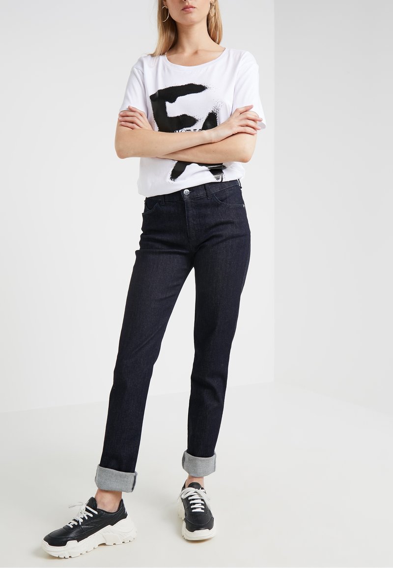 Camiseta blanca con un diseño gráfico en negro, combinada con jeans de mezclilla oscura y zapatillas negras. Los jeans tienen los puños doblados y un corte clásico.