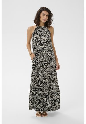 Kobieta o kręconych włosach, ubrana w czarno-białą długą sukienkę w paisley bez rękawów i sandały, stojąca z jedną ręką w kieszeni.