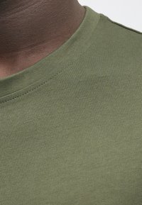 Olivově zelené bavlněné tričko s kulatým výstřihem. Vyznačuje se hladkou, mírně texturovanou látkou a jednoduchým, klasickým designem bez ozdob.