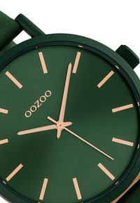 Montre verte avec une finition mate, dotée d'index et d'aiguilles dorées, ainsi que d'un bracelet vert texturé. Nom de la marque "OOZOO" affiché.