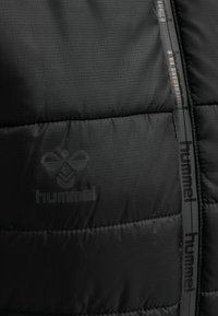 Svart vadderad jacka med rutig mönstring, med en diskret präglad logotyp och märkestejp längs dragkedjan. Slät textur.