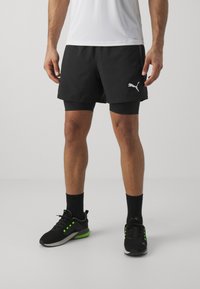 Zwarte sportshorts met een binnenvoering, voorzien van een wit Puma-logo. Gecombineerd met zwarte sneakers met groene accenten.