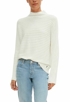 Pull à col roulé blanc côtelé à manches longues associé à un jean taille haute bleu clair porté par une personne debout.