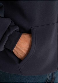 Hand resting inside the front pocket of a dark navy sweatshirt with ribbed cuff, paired with blue denim jeans.