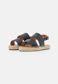 Sandali neri in pelle con due ampie cinghie e chiusura a fibbia, dotati di suole in stile espadrilles intrecciate su uno sfondo bianco.