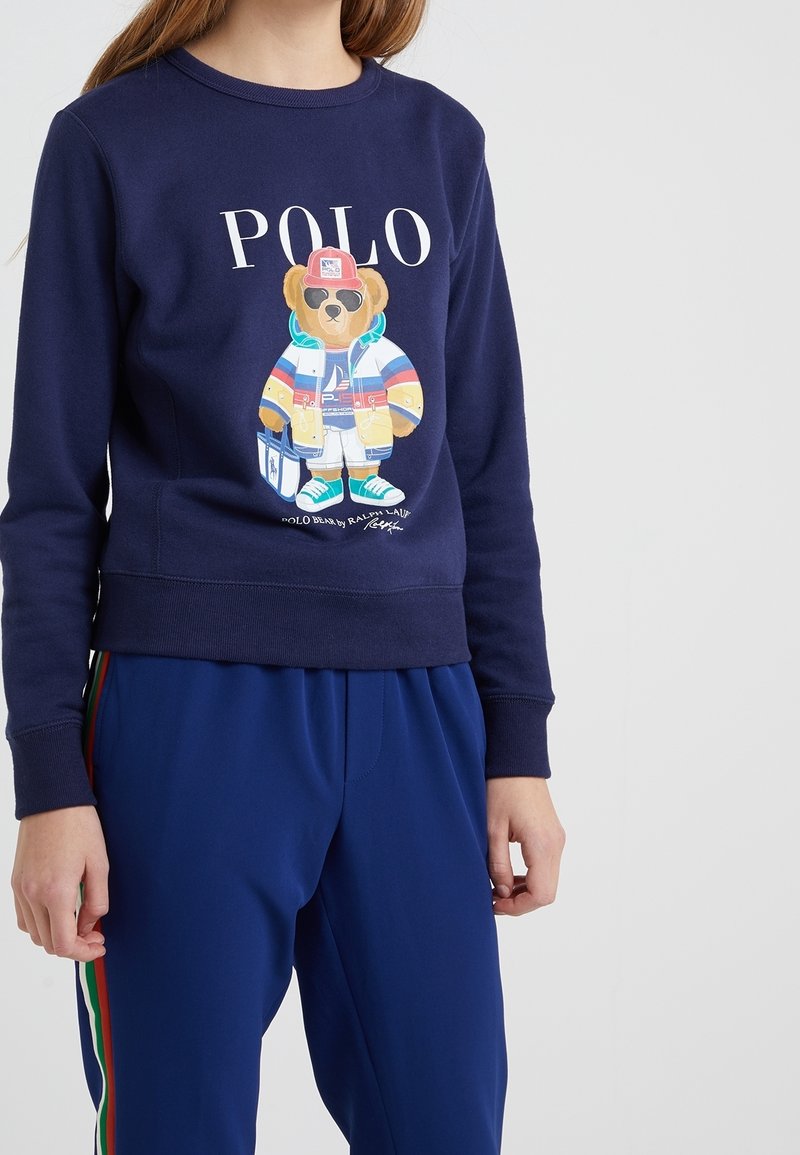 Felpa navy con una grafica di un orso vestito in abbigliamento colorato. Polsini e orlo a costine. Abbinata a pantaloni blu con strisce colorate.