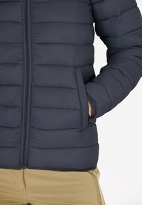 Marinblå puffjacka med horisontell quiltad design, dragkedjeslutning framtill och sidofickor. Mjukt, lätt material med en slät yta.